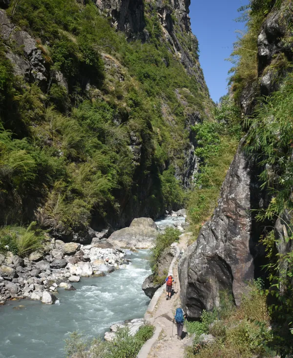 Hikers on a narrow path carved into rock above a blue river rushing through a steep canyon