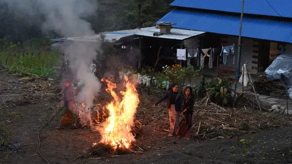 Villagers gathered around a fire outside a blue-roofed house at dusk