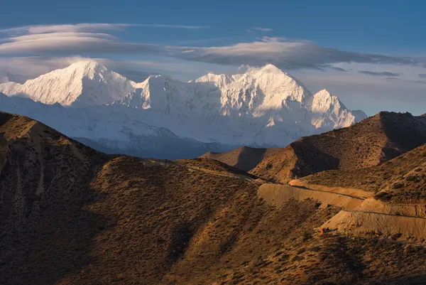 Late sunlight catching rugged brown foothills as clouds swirl around glowing Himalayan peaks