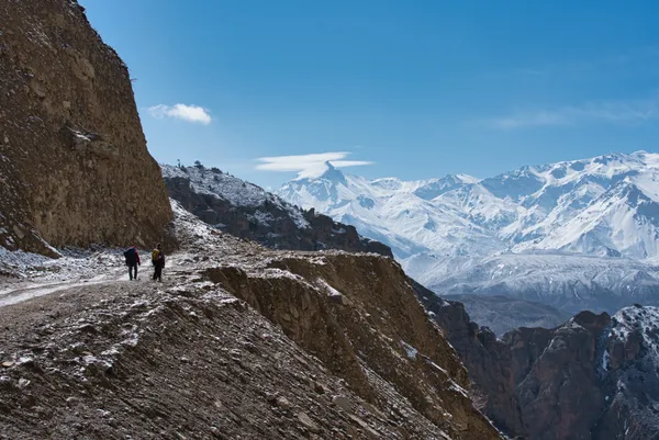 Hikers traversing a narrow mountain road overlooking deep canyons and distant Himalayan peaks