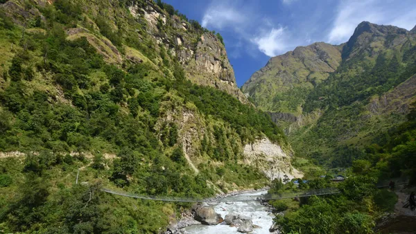 A metal suspension bridge spanning a rushing river between green hills