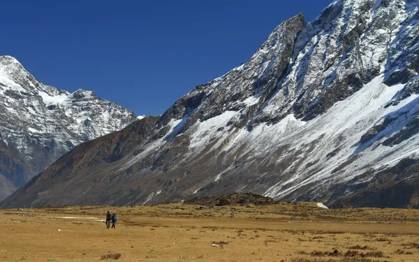 Two hikers traversing vast barren landscape beneath steep snow-dusted slopes