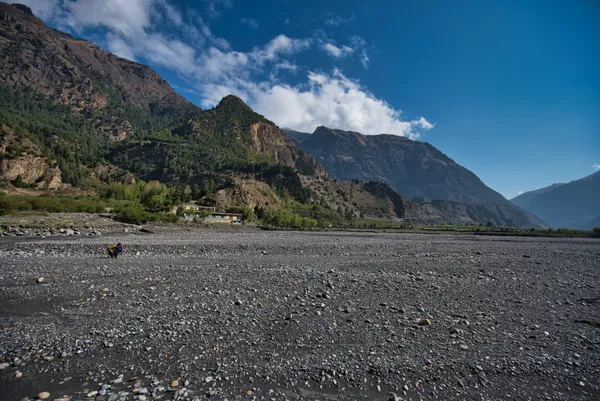 A lone traveler crossing an expansive pebble-strewn riverbed beneath towering rocky peaks
