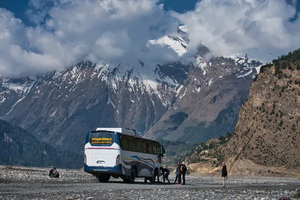 A local bus paused on a valley floor beneath cloud-draped snowy mountain summits