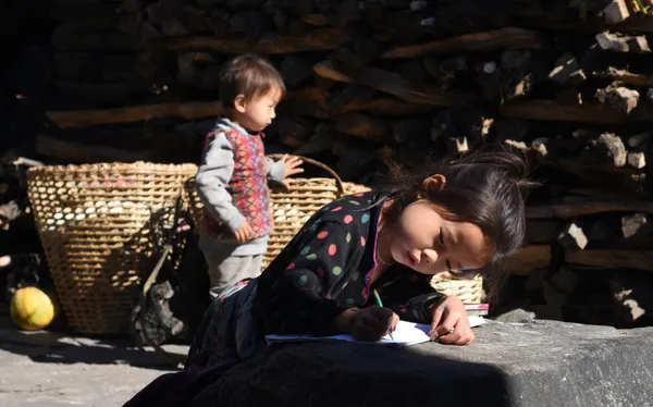 A girl concentrating on schoolwork while a young boy plays nearby in a village courtyard