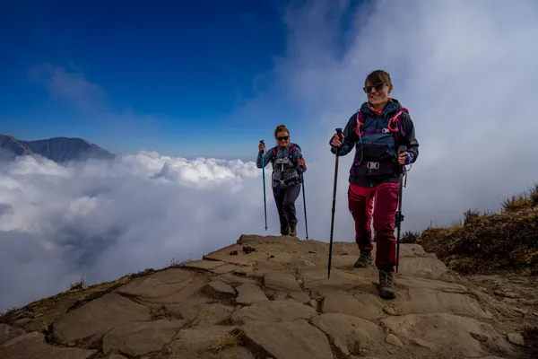 Two trekkers walking on a stone ridge path above the clouds
