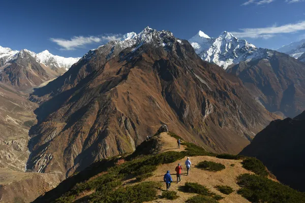 A group on a narrow ridge with snow-capped peaks rising behind