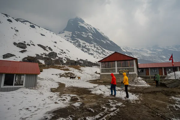Two trekkers beside red-roofed lodges surrounded by snow at base camp