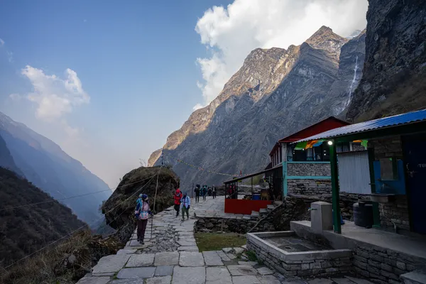 Hikers on a stone-paved path beside a blue-roofed teahouse below cliffs