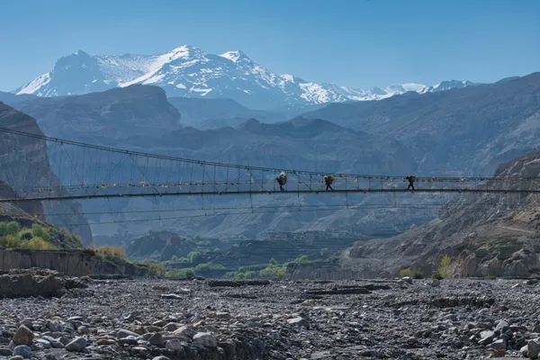 Porters crossing a high suspension bridge over the stony riverbed toward Tangya Village at 3,240 m