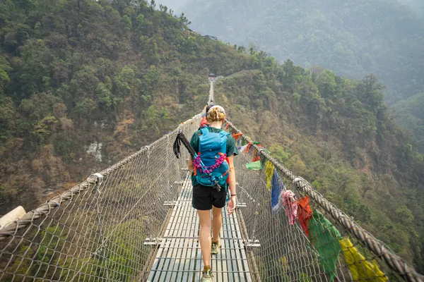 A trekker with a blue backpack walking across a suspension bridge