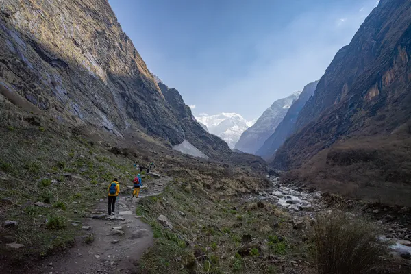 Hikers following a rocky river valley toward distant snowy peaks